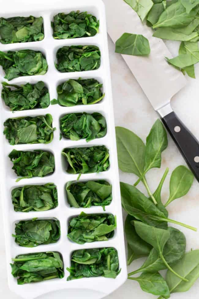 A white ice cube tray filled with green vegetable leaves. A knife with black handle sits next to the ice cube tray.