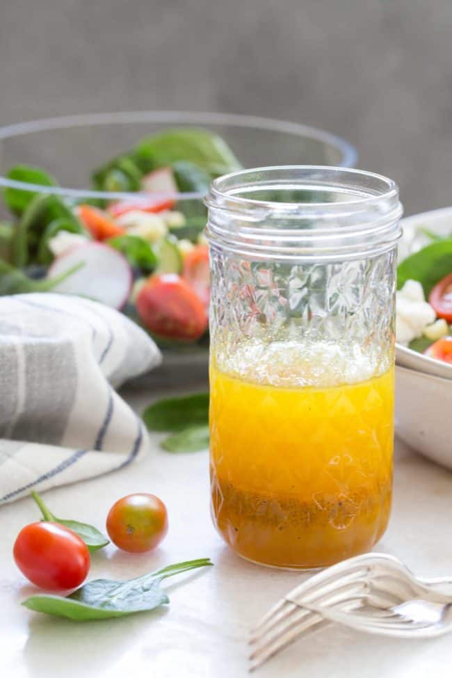 Glass mason jar with a spoon in it and half filled with avocado oil salad dressing. A white bowl of salad and two forks are next to the mason jar.