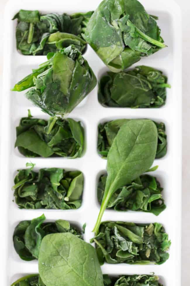 A white ice cube tray filled with frozen spinach leaves