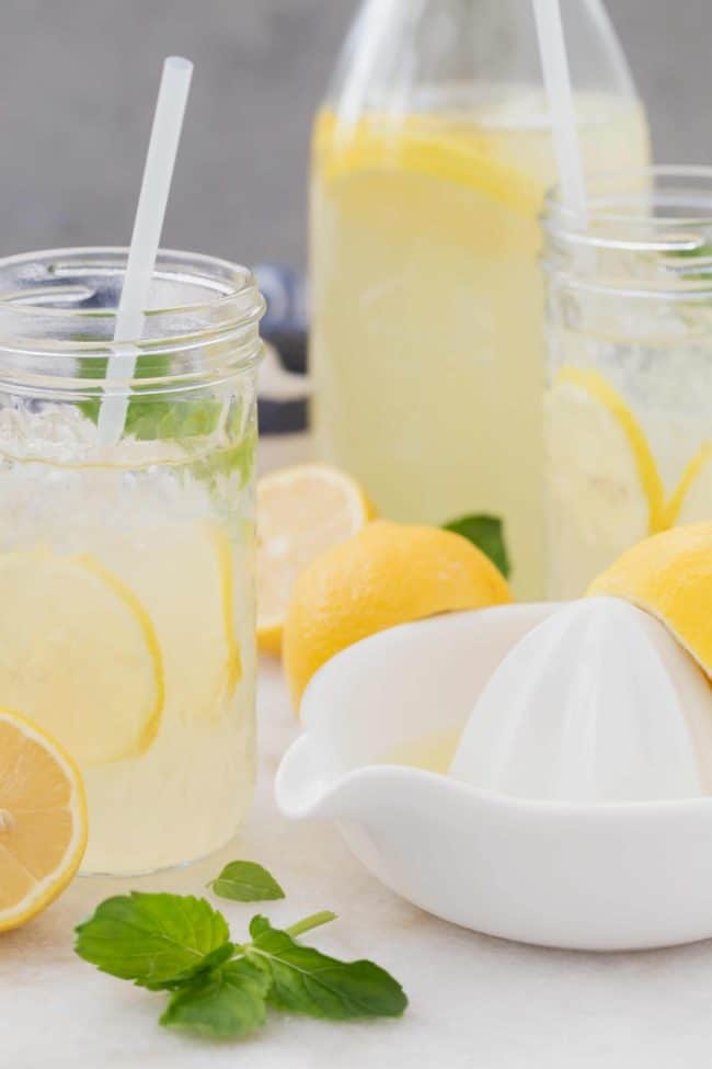 Glass jars filled with lemonade with honey. A white juicer with lemons sits next to the glasses to make a lemonade recipe with honey..