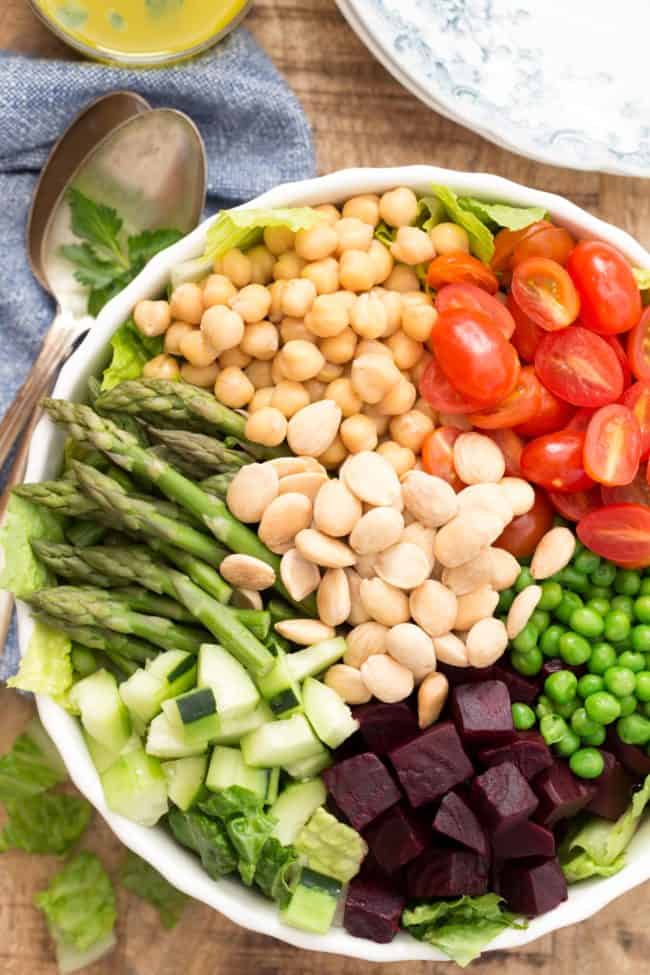 White bowl filled with asparagus, chopped beets, peas, cherry tomatoes, lettuce and garbanzo beans. Two plates and two spoons sit next to the bowl.for salad.