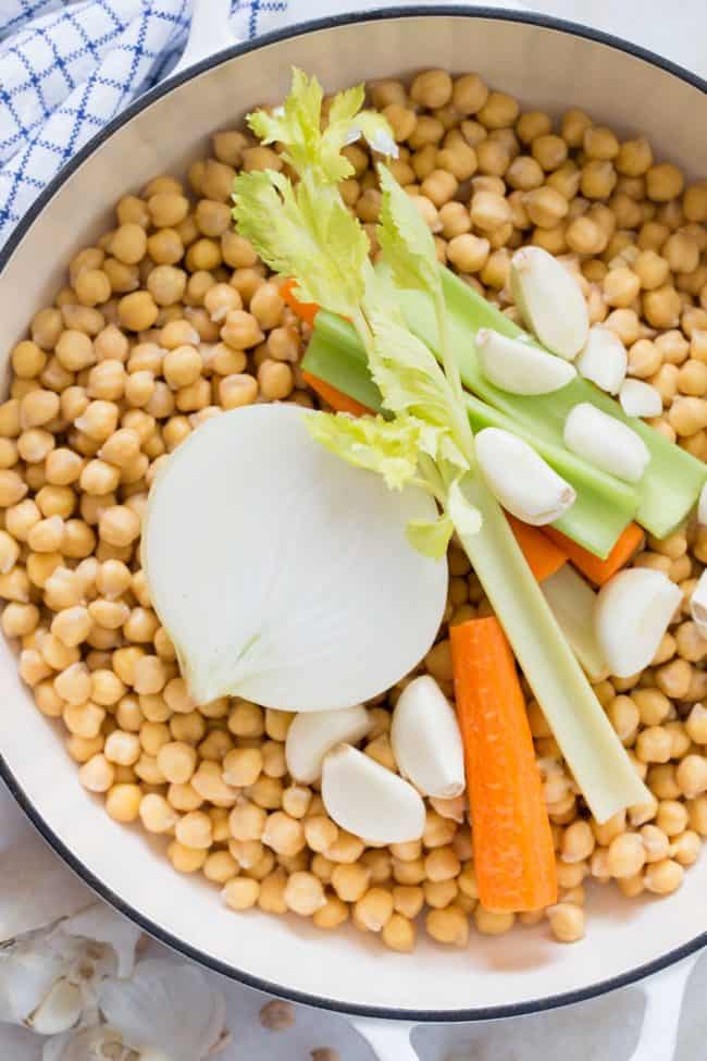 A white pot filled with legumes, celery stalks, garlic cloves, carrot pieces and an onion half. - For Are chickpeas gluten free