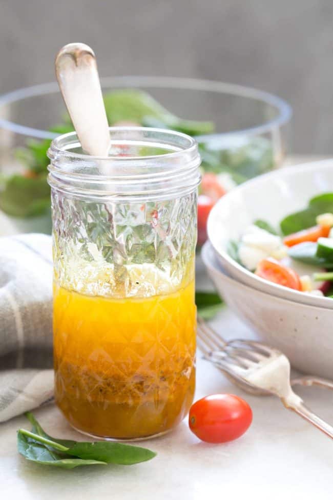 Glass mason jar with a spoon in it and half filled with vinaigrette. A white bowl of salad and two forks are next to the mason jar.