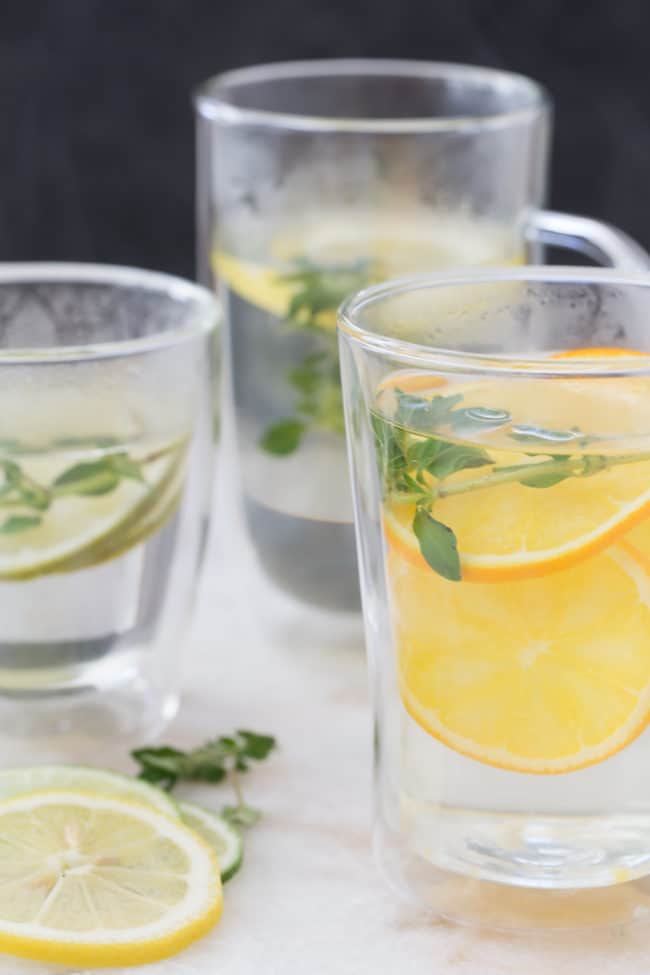 Three clear cups of hot water with fresh herbs and fruit slices for herbal oregano tea recipe