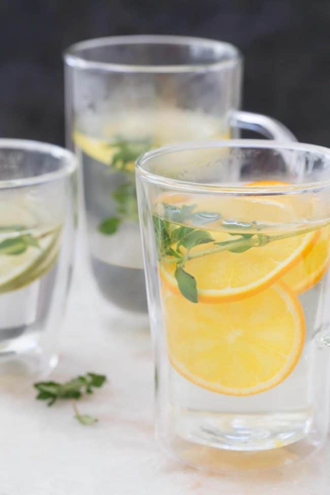 Two clear cups of hot water with fresh herbs and fruit slices.