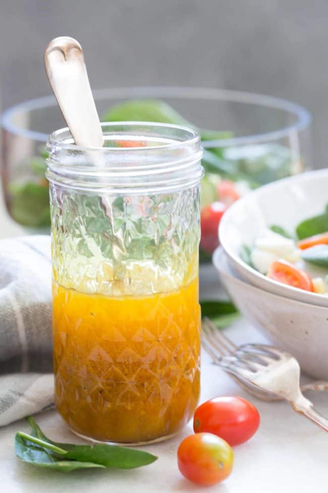 Glass mason jar with a spoon in it and half filled with avocado oil salad dressing. A white bowl of salad and two forks are next to the mason jar.