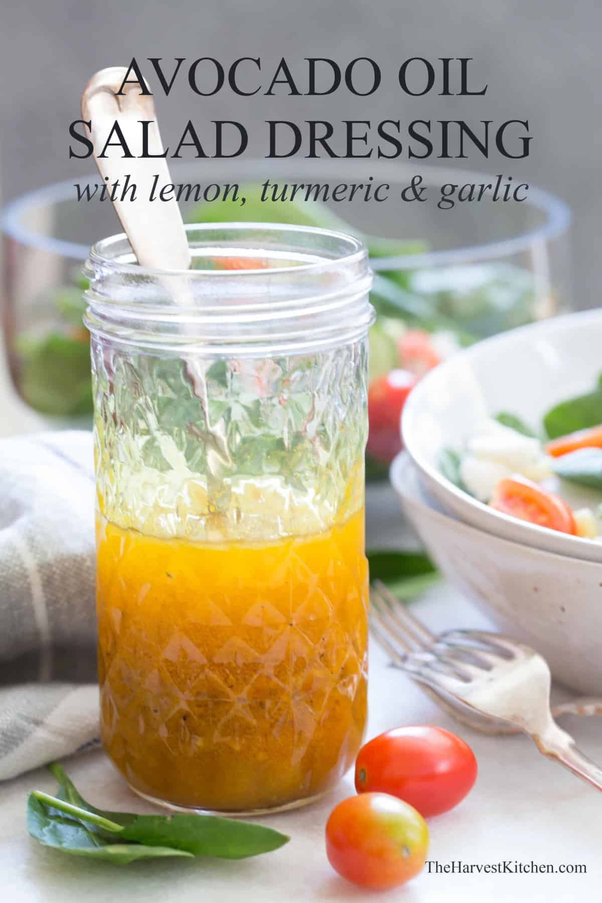 Glass mason jar with a spoon in it and half filled with avocado oil salad dressing. A white bowl of salad and two forks are next to the mason jar.