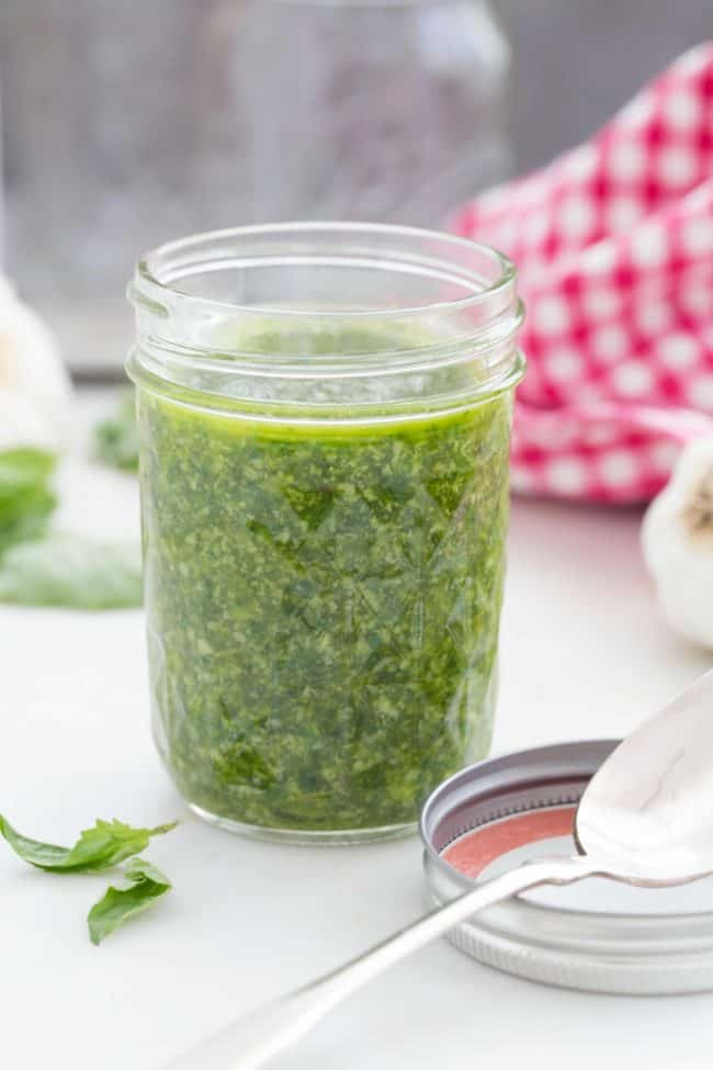 A clear glass jar filled with homemade basil pesto. A spoon sits next to the jar.