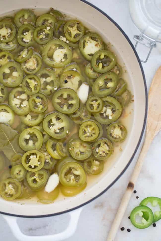 A white Dutch oven filled with Pickled Jalapenos. A wooden spoon rests next to the pot.