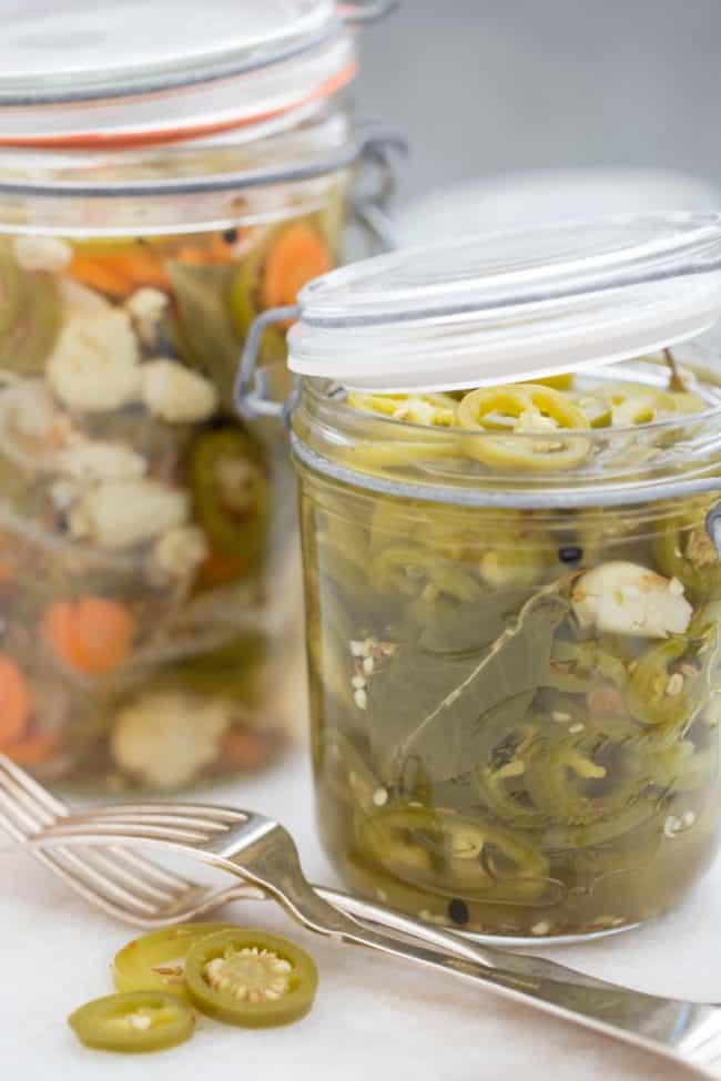 Two clear glass mason jars filled with sliced pickled jalapenos. Two forks sit next to the jars.