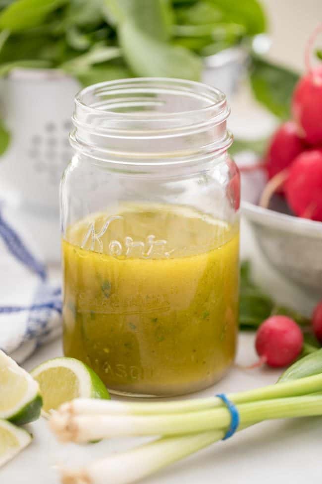 A glass mason jar half filled with homemade lime vinaigrette. A white colander in the back filled with lettuce.