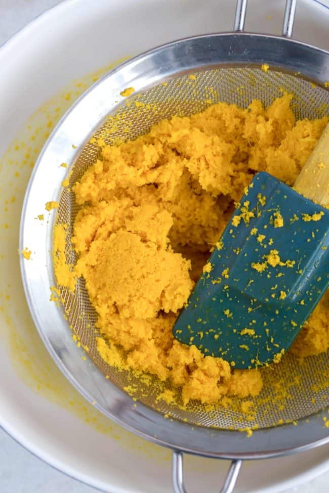 Blended root juice being pressed through a fine mesh sieve into a white bowl.