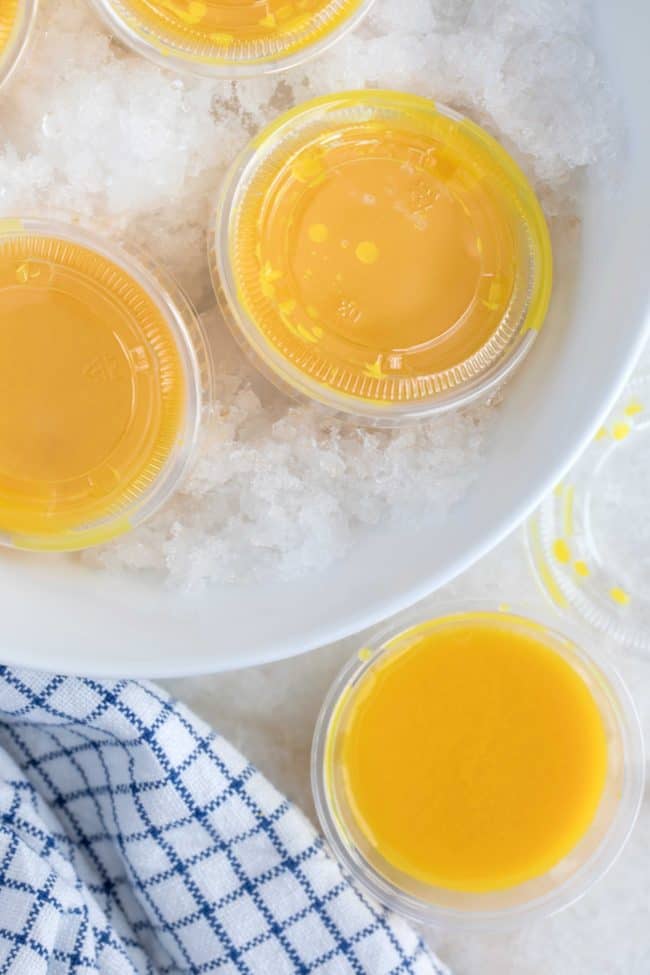 Five small cups filled with ginger turmeric shots sit in a white bowl filled with crushed ice.