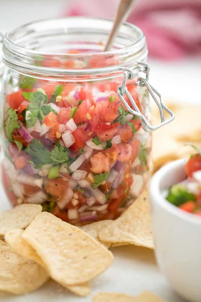 A clear glass mason jar filled with Mexican salsa fresca (Pico de Gallo). A spoon is in the jar and corn tortilla chips are scattered around the jar.