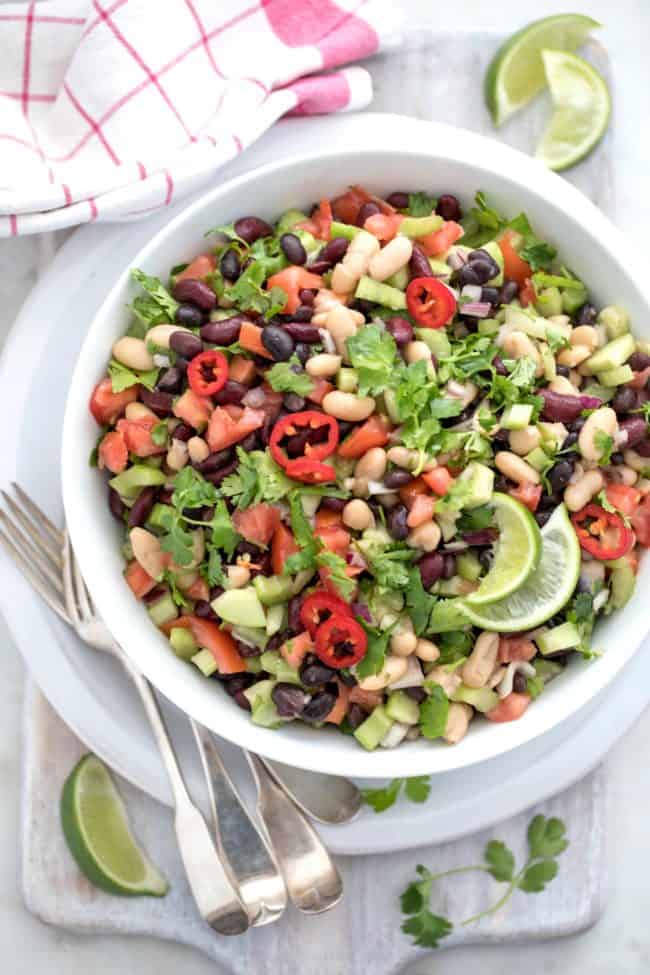 A white bowl filled with tossed beans and vegetables. Red and white dish towel next to the bowl with a serving spoon. 