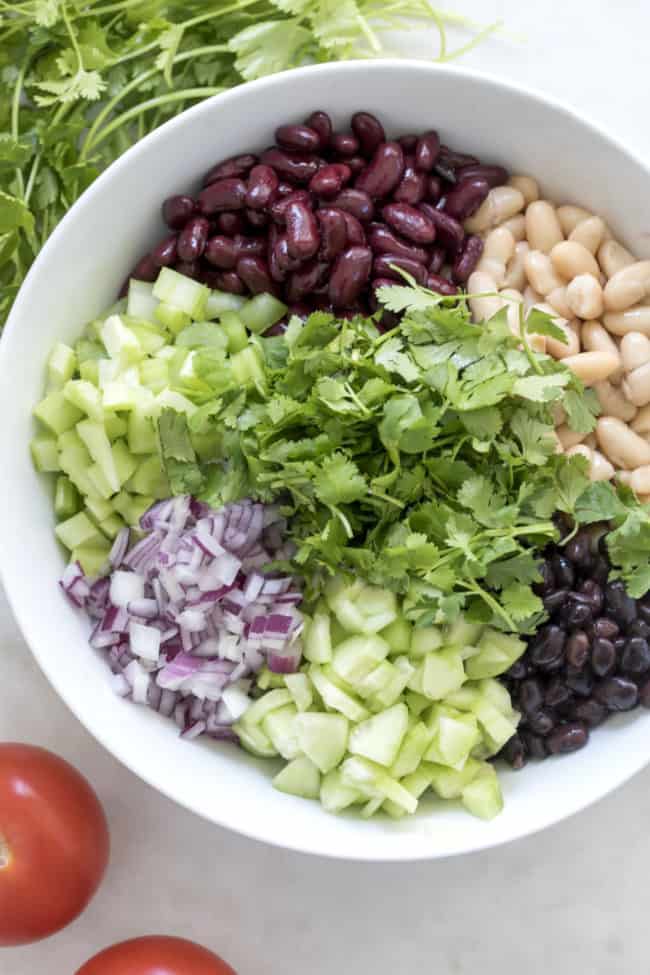 A white bowl of legumes, chopped onion, chopped cucumber and cilantro.