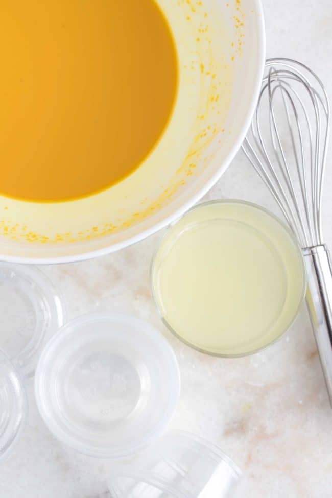 A white bowl filled with ground turmeric juice to make lemon ginger and turmeric shots. A clear glass filled with lemon juice sits next to the white bowl.