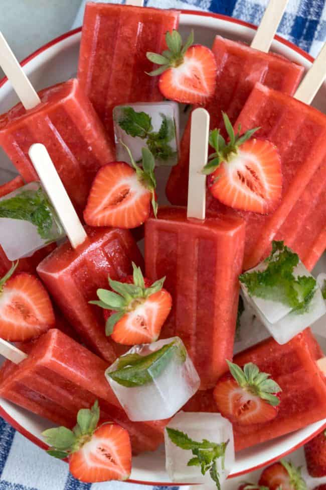 A white bowl filled with strawberry popsicles Fresh strawberries are scattered over the popsicles.