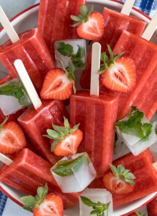 A white bowl filled with ice and strawberry popsicles. Fresh strawberries are scattered over the popsicles.