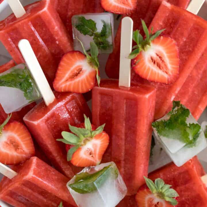 A white bowl filled with ice and strawberry popsicles. Fresh strawberries are scattered over the popsicles.