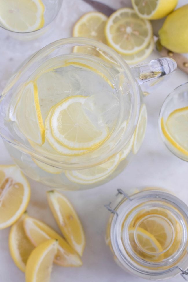 A glass pitcher filled with water and lemon slices. Lemon slices with a knife sit next to the pitcher.