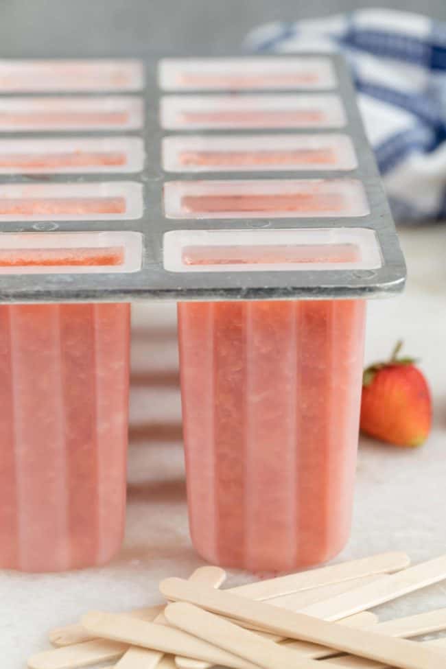 A plastic popsicle mold filled with pink juice. Wooden popsicle sticks are scattered next to the plastic mold.