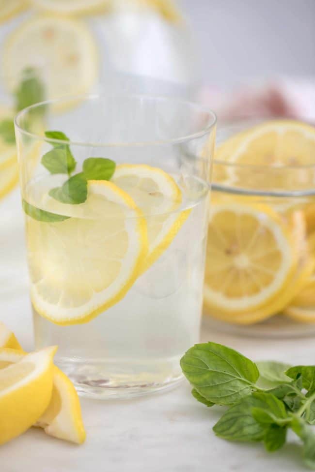 Two clear drinking glasses filled with lemon water. Slices of lemon and sprigs of mint are in the glasses..