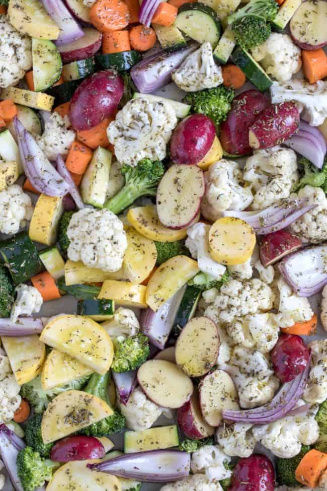 A cookie sheet filled with raw cauliflower, potatoes, zucchini, carrots and onions with seasoning sprinkled on them