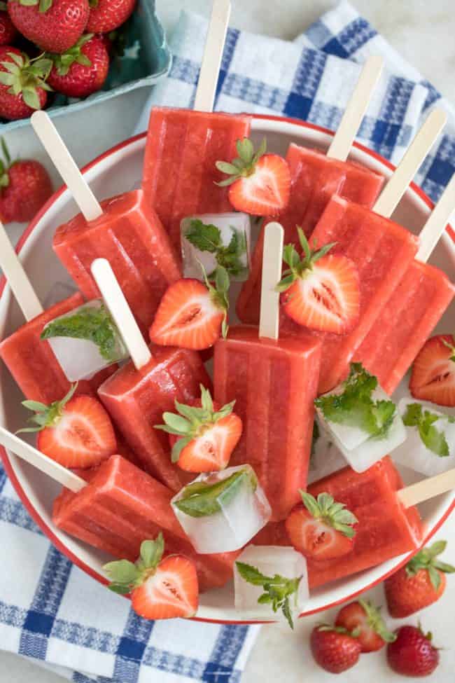 A white bowl filled with ice and strawberry popsicles. Fresh strawberries are scattered over the popsicles.