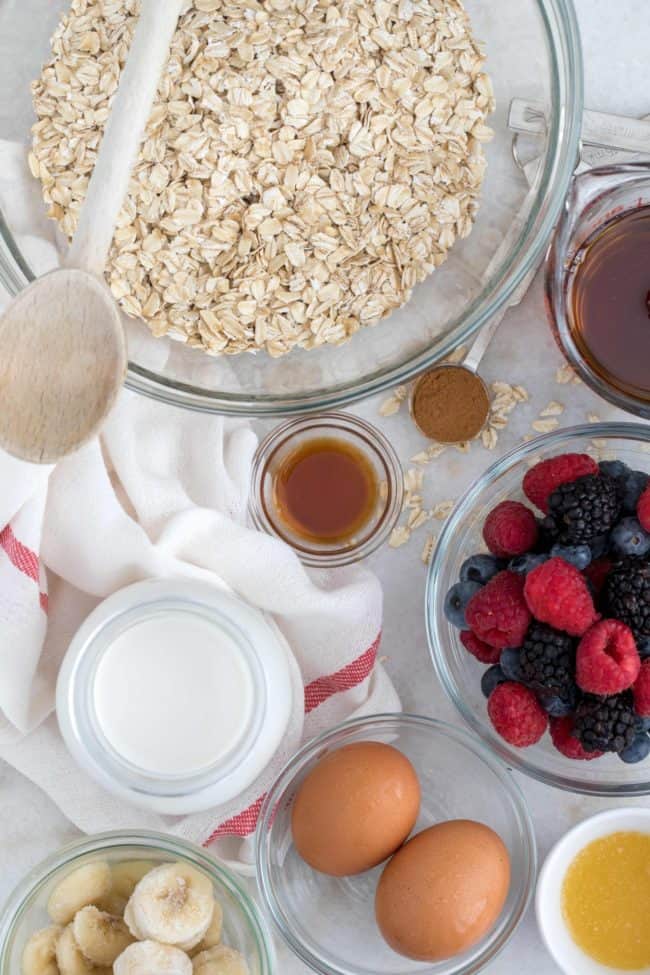 Clear glass mixing bowls filled with oats, milk, eggs, banana and berries. 
