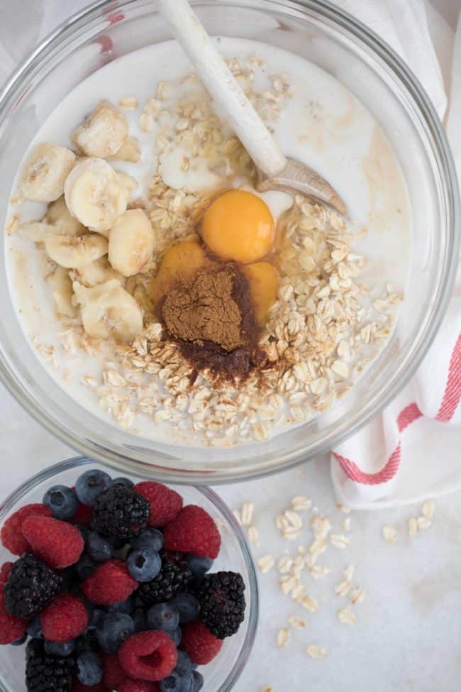 Two clear glass mixing bowls filled with oats, raw eggs, banana and the other bowl if filled with berries.