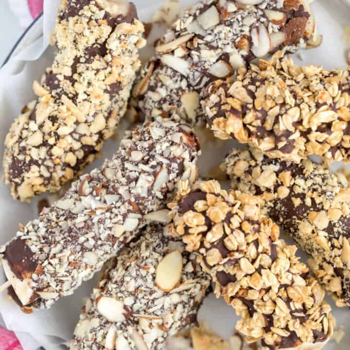 A white plate filled with Frozen Chocolate Covered Bananas are frozen bananas dipped in dark chocolate then rolled in chopped nuts, granola and shredded coconut