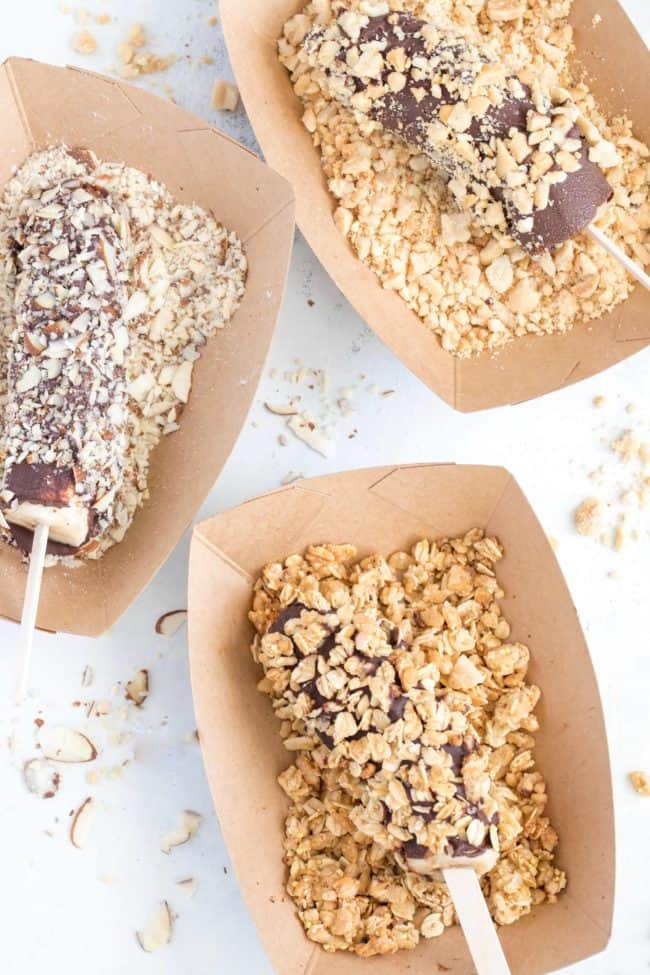 Three frozen chocolate covered bananas dipped in chopped nuts and granola.