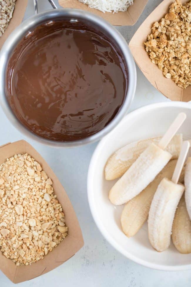 Frozen fruit on a stick in a bowl next to bowls of a creamy dark sauce and chopped nuts.
