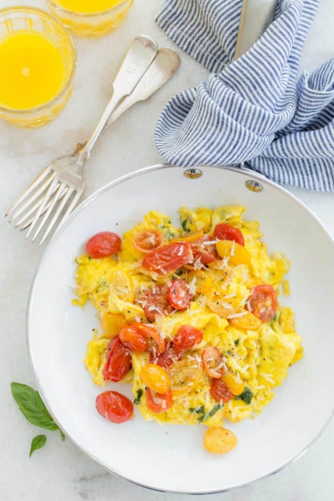 A white skillet filled with scrambled eggs with tomatoes, basil and Parmesan cheese.