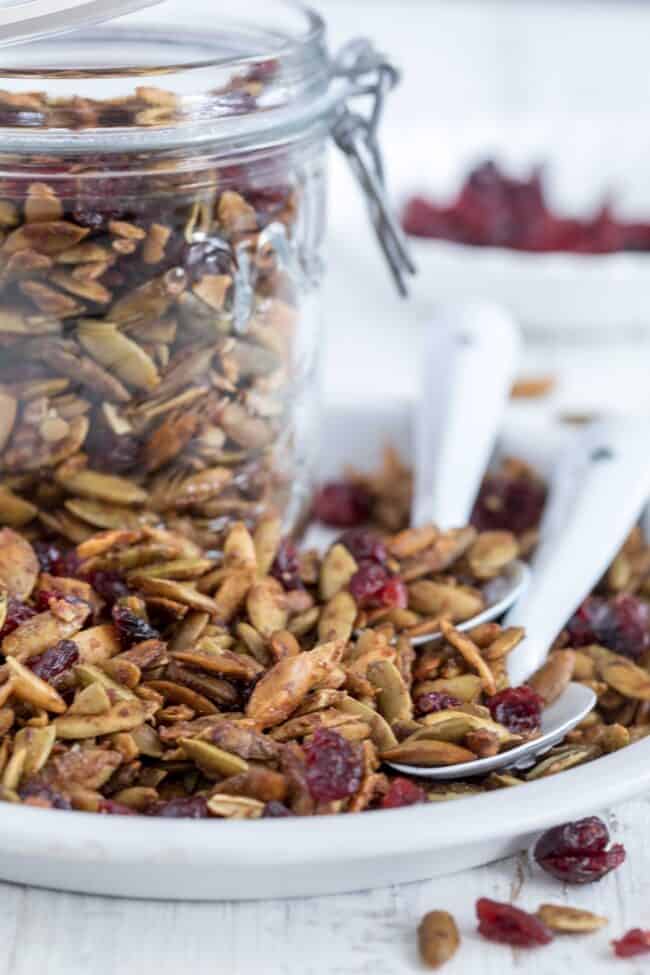 A mason jar filled with roasted pumpkin seeds and dried cranberries. The mason jar sits on a white dish with two white spoons.