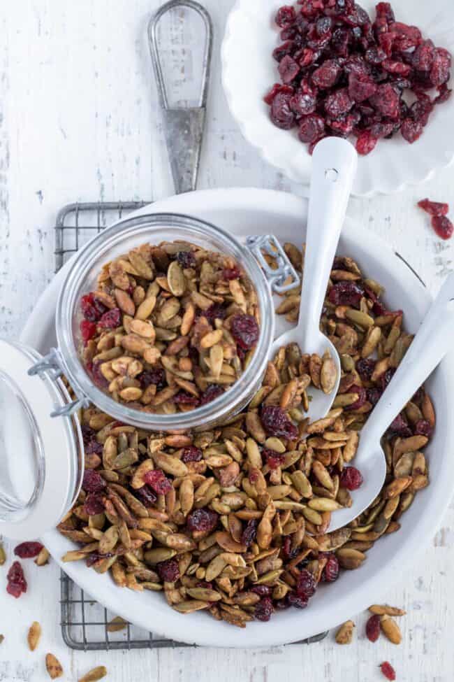 A white dish filled with roasted pumpkin seeds and dried cranberries. Two white spoons are tucked in the seeds.Roasted Pumpkin Seeds in dish