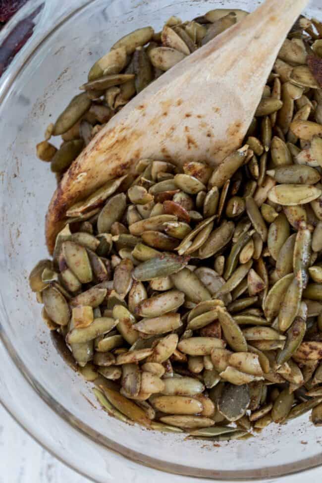 A clear bowl filled with raw pepitas. A wooden spoon sits in the bowl.