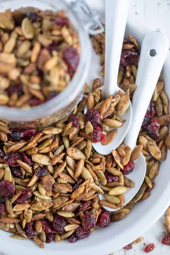 A white dish filled with roasted pumpkin seeds and dried cranberries. Two white spoons are tucked in the seeds.Roasted Pumpkin Seeds in dish