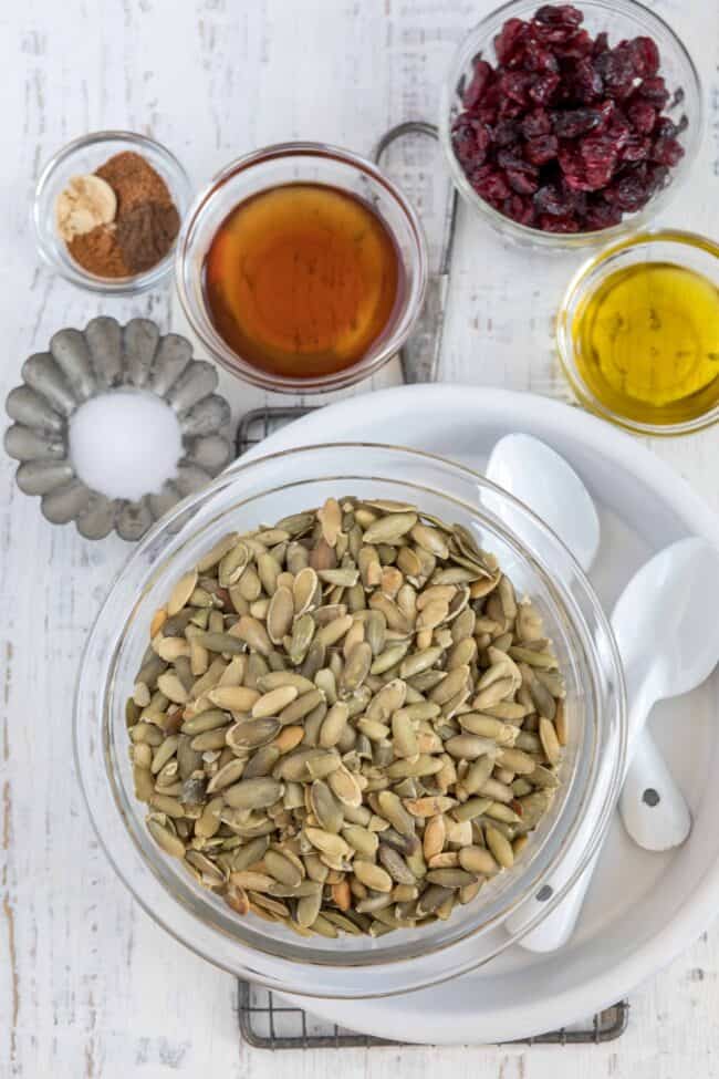 A clear bowl filled with seeds. Small glass bowls next to it filled with seasoning, maple syrup, dried cranberries and oil.