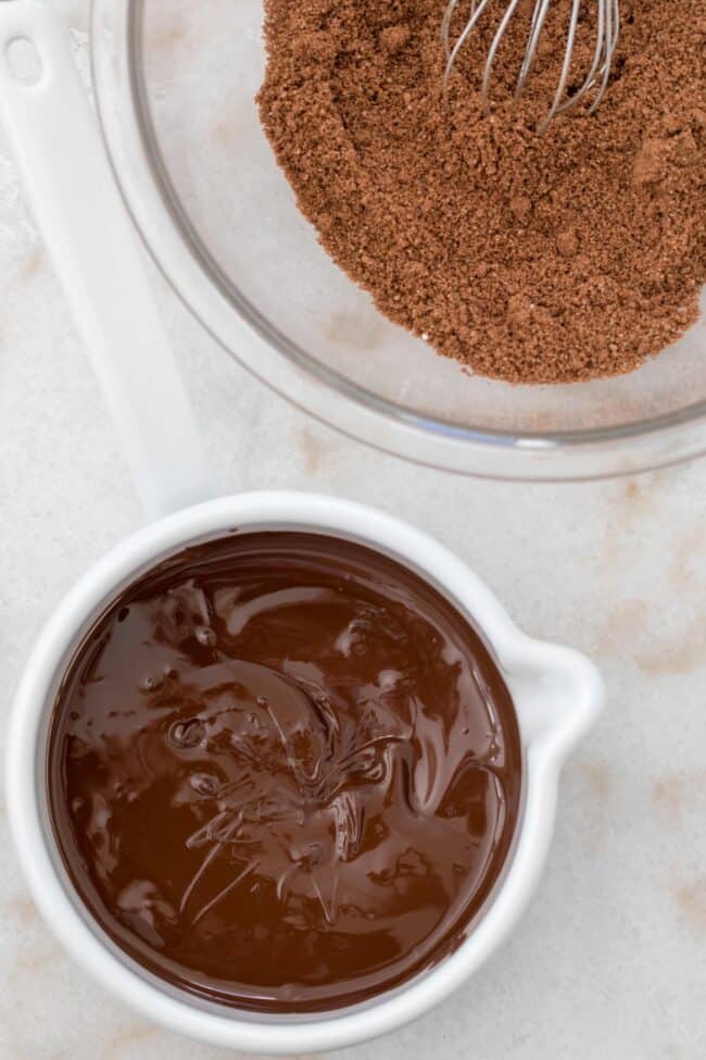 A glass bowl filled with almond flour and a small white pot filled with melted chocolate for almond flour brownies.
