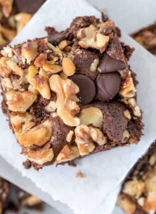 A chocolate brownie with walnuts on top sits on white parchment paper.