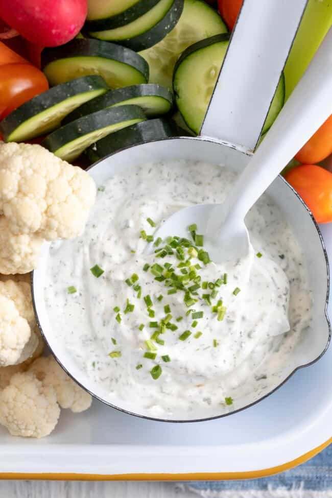 A white bowl filled with ranch dip. An assortment of vegetables are scattered around the dip.