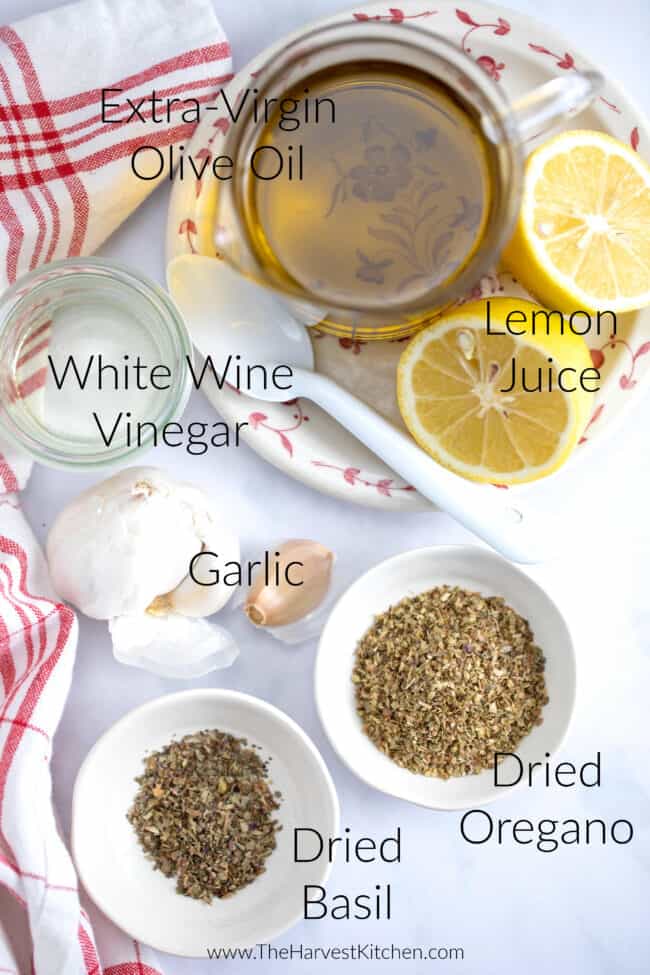 glass bowls filled with oregano, basil, white wine vinegar and olive oil. A white and red dish towel sits next to the bowls.