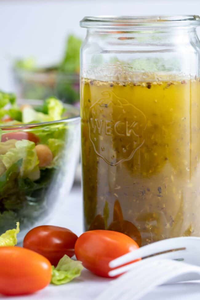 glass jar filled with Italian salad dressing. Clear glass bowl filled with salad and two forks and cherry tomatoes lay next to mason jar.