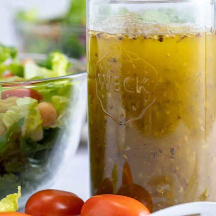 glass jar filled with Italian salad dressing. Clear glass bowl filled with salad and two forks and cherry tomatoes lay next to mason jar.