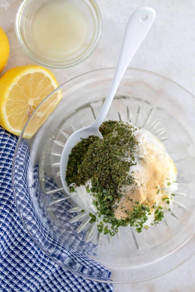 A clear glass bowl filled with mayonnaise, yogurt and seasoning. A white mixing spoon rests in the bowl.