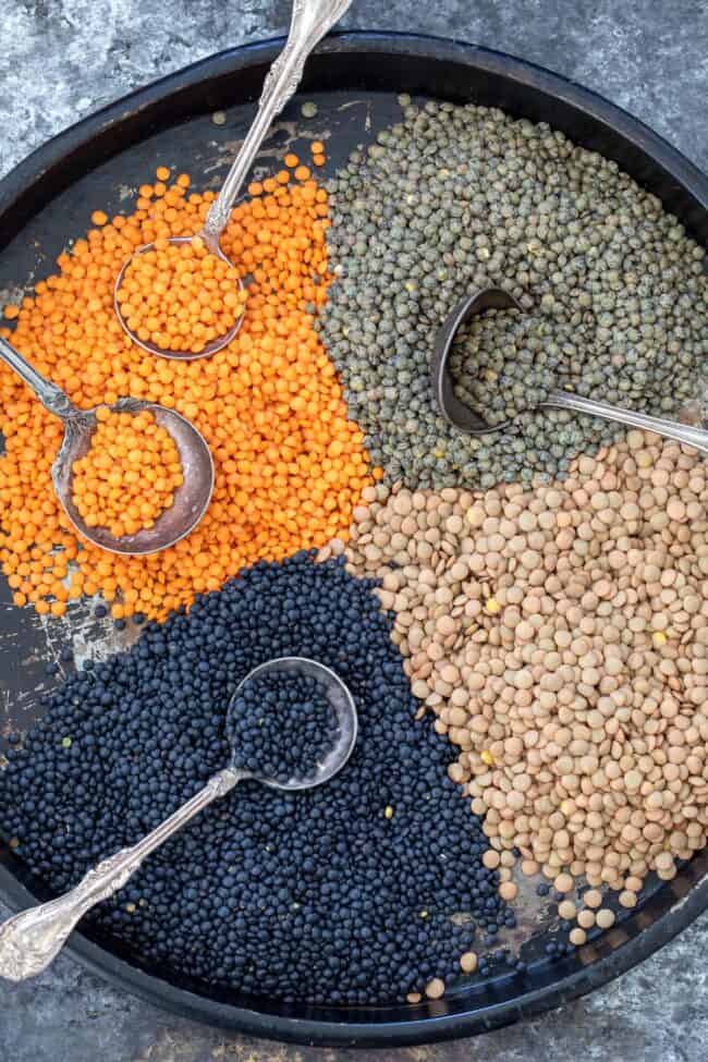 A black tray with different types of dried lentils scattered on it. Four spoons rest in the lentils.