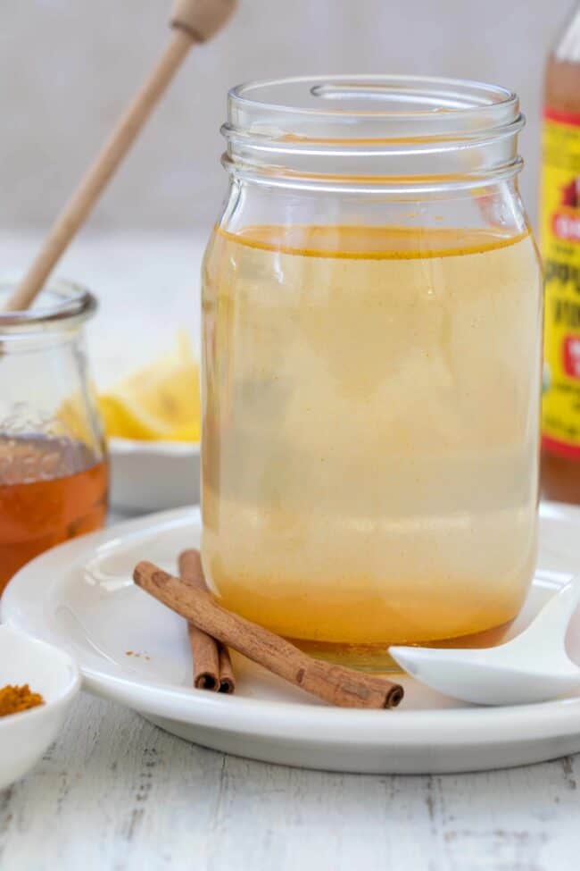 A clear glass mason jar filled with Bragg ACV and lemon juice and water. A cup of honey sits next to it.