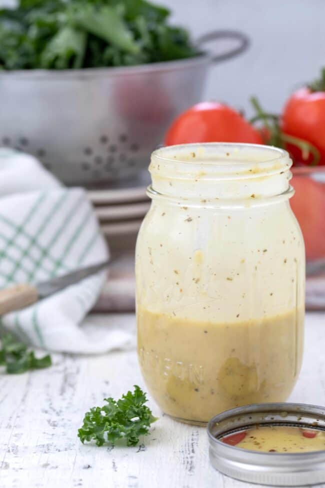 A clear mason jar with homemade apple cider vinegar salad dressing. A metal colander filled with lettuce and tomatoes in the background.